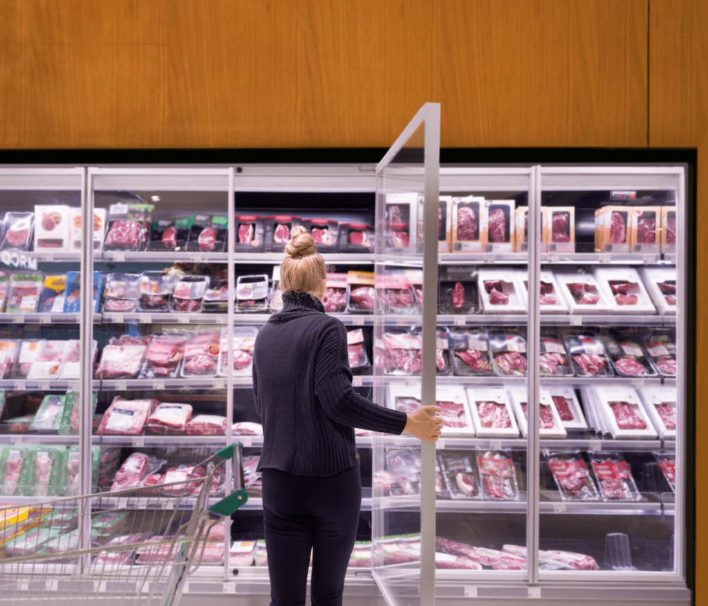 A person standing in a supermarket looking at animal meat products in a fridge