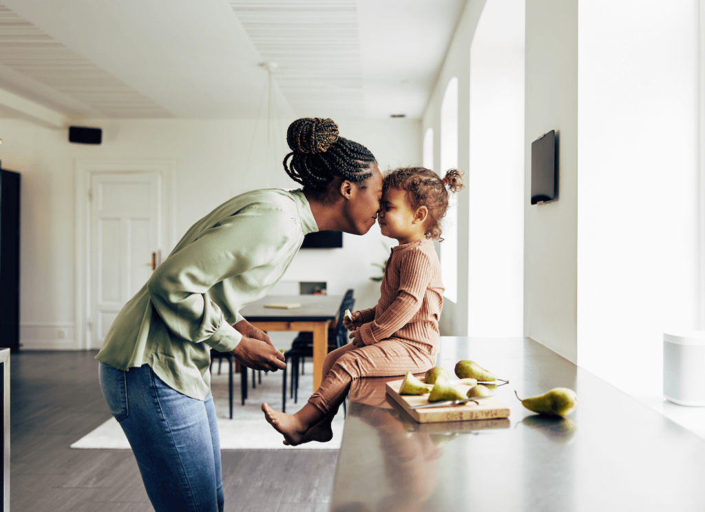 A parent interacts with their child, who is sitting on a kitchen bench beside some chopped fruit