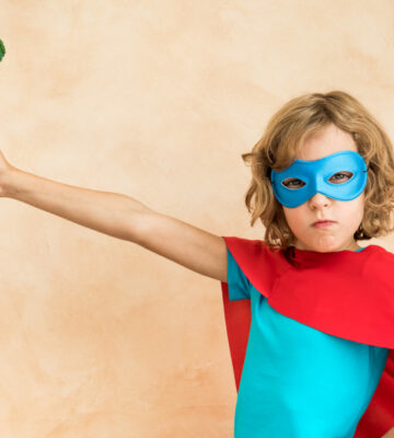 A child dressed as a superhero holding a piece of broccoli on a fork