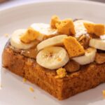 A slice of vegan banana bread topped with plant-based honeycomb, nuts, and slices of banana