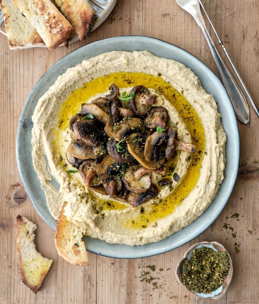 A dish of freshly prepared vegan creamy mushroom hummus with olive oil drizzled on top and crusty bread surrounding the bowl
