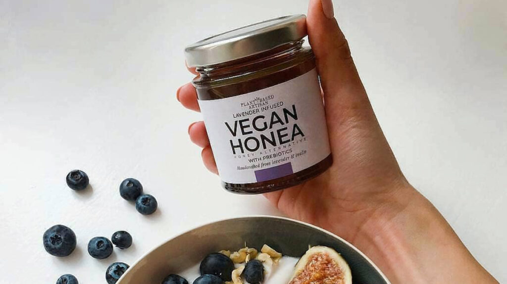 A hand is holding a jar of Plant Based Artisan Vegan Honea, above a bowl of blueberries