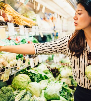 A young asian woman reaches for some vegetables in a supermarket. She is holding a lettuce.