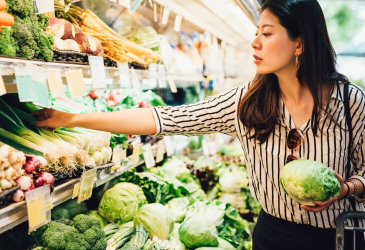 A young asian woman reaches for some vegetables in a supermarket. She is holding a lettuce.