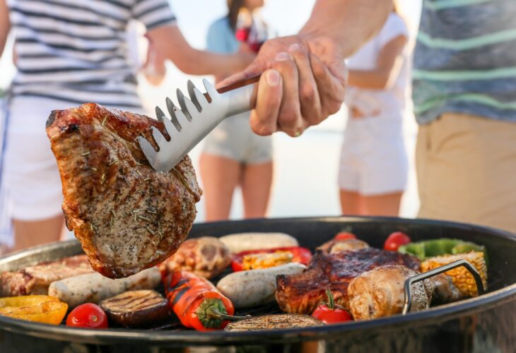 A person holds steak with metal tongs over a barbecue