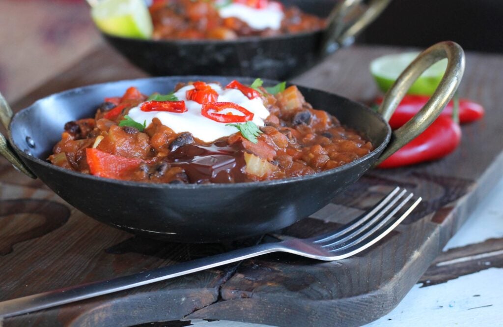 A bowl of vegan chilli hotpot made with chocolate, in a black bowl, topped with vegan sour cream and fresh chillis, with a fork beside it