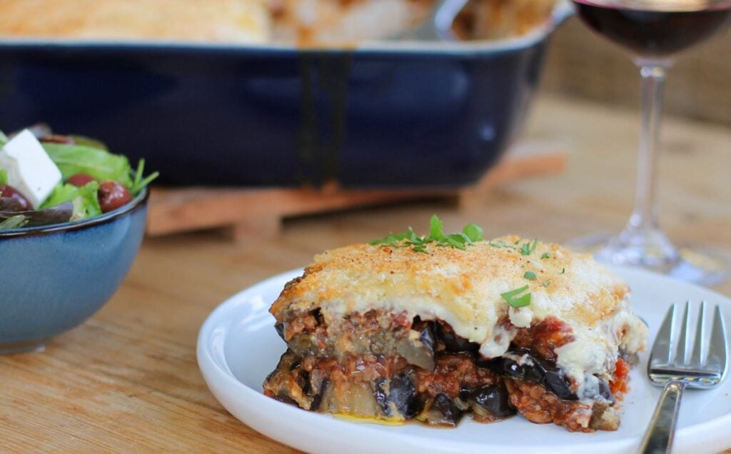A square portion of Greek moussaka made to a vegan recipe, presented on a white circular plate with a silver fork