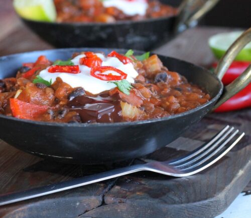 A bowl of vegan chilli hotpot made with chocolate, in a black bowl, topped with vegan sour cream and fresh chillis, with a fork beside it