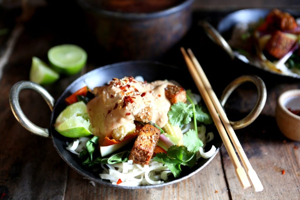 A wok filled with a vegan starry, topped with peanut satay sauce, with chopsticks to the side