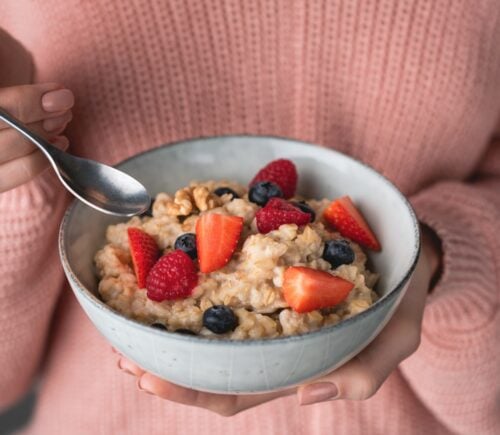 A person wearing a pink jumper holds a bowl of fruit and oatmeal