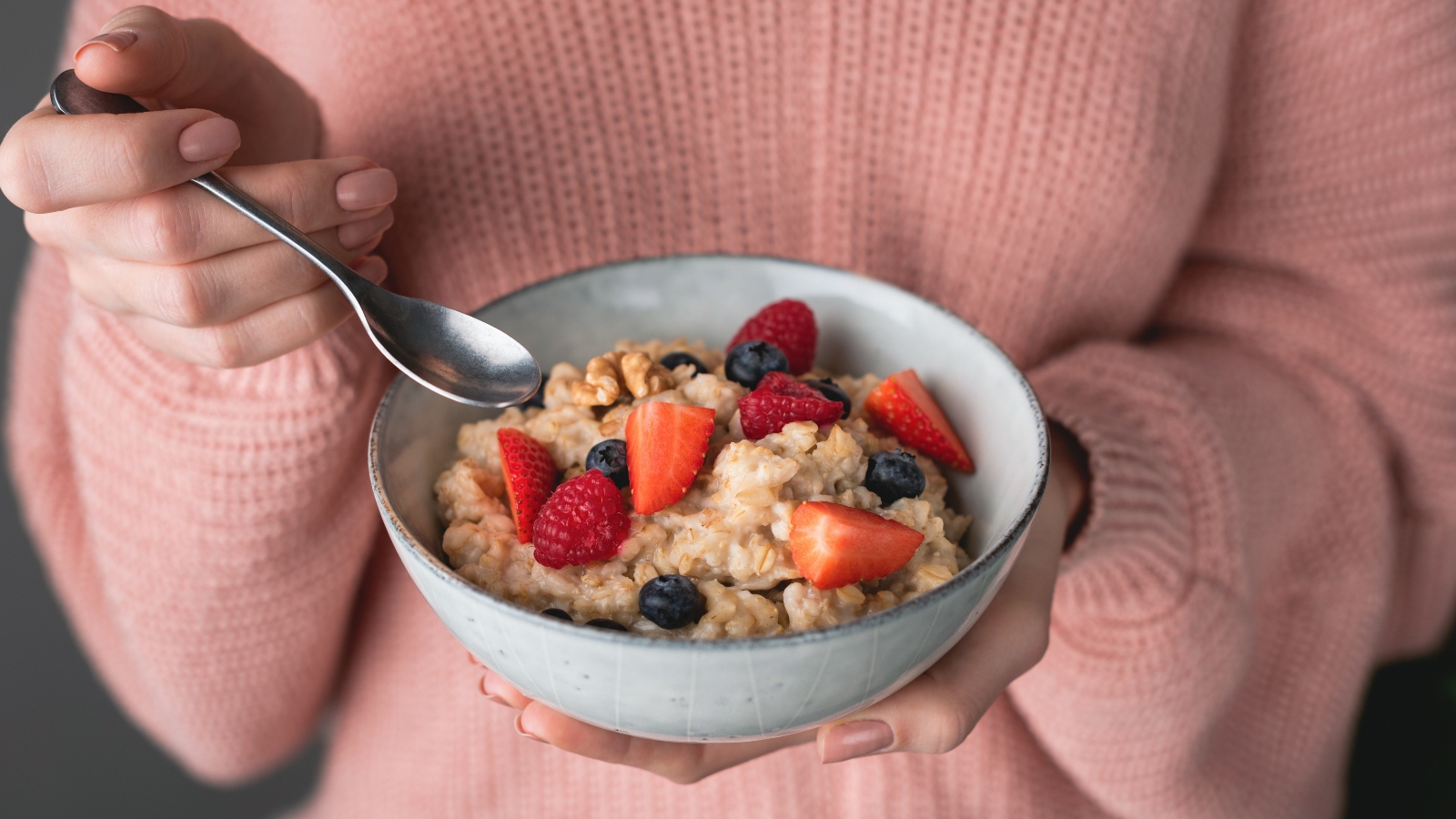 A person wearing a pink jumper holds a bowl of fruit and oatmeal