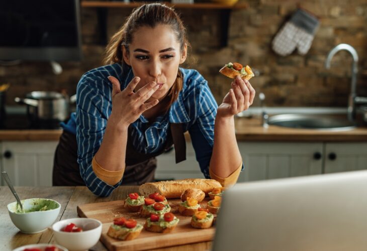 A woman eats avocado and tomato on bread while looking at a laptop