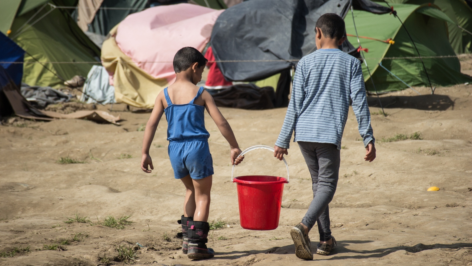 Two young children carry a bucket of water in front of tents