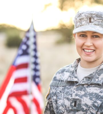A woman dressed in army uniform stands next to a US flag