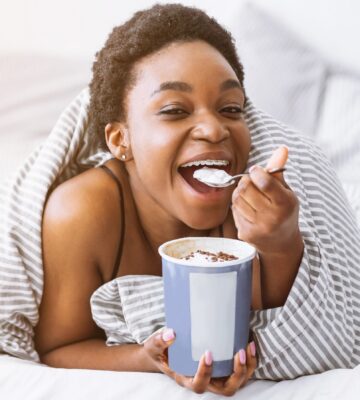 A woman lies on a bed wrapped in a duvet eating ice cream