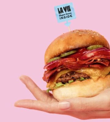 A person holds a vegan bacon burger against a pink background