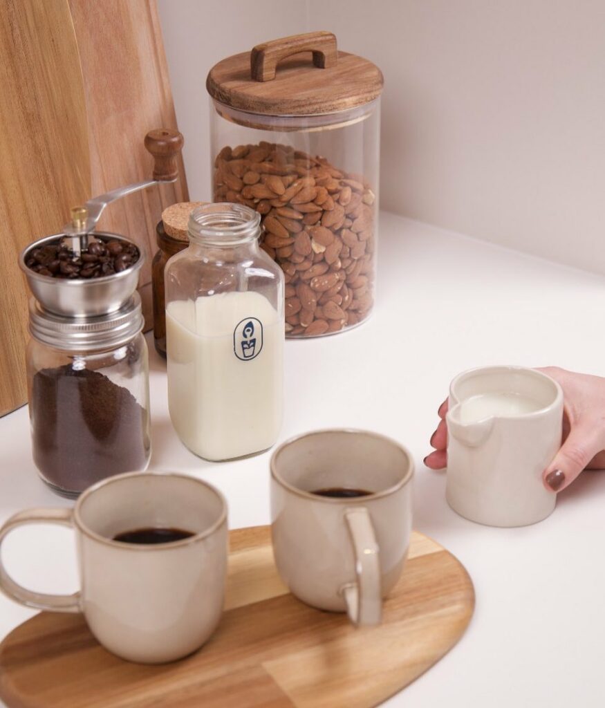 Mugs, a bottle of vegan milk, and almonds in a jar on a kitchen bench