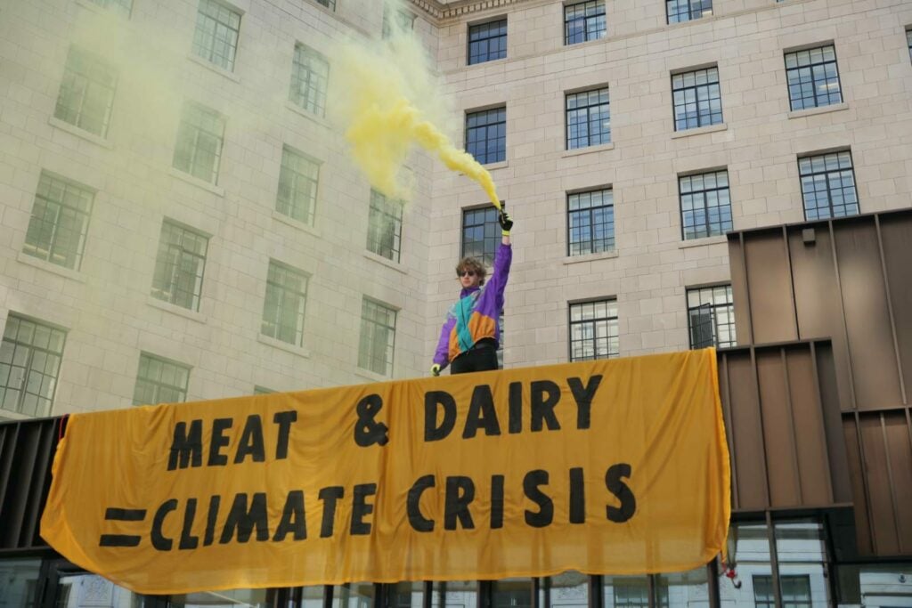 A person holds a smoke bomb above a sign that says "meat and dairy climate crisis" outside a university