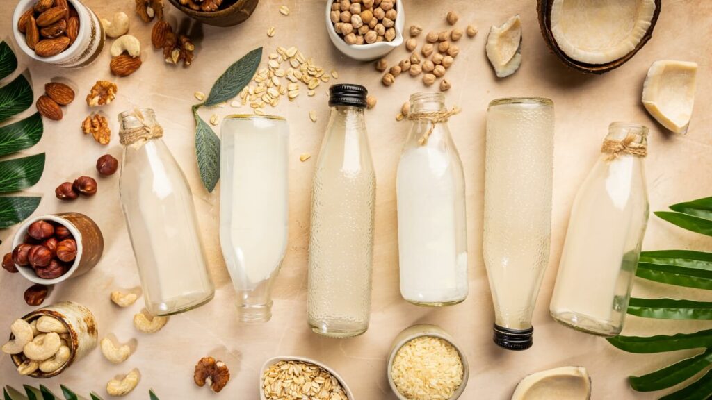 Six bottles of vegan milk lying down among nuts and seeds
