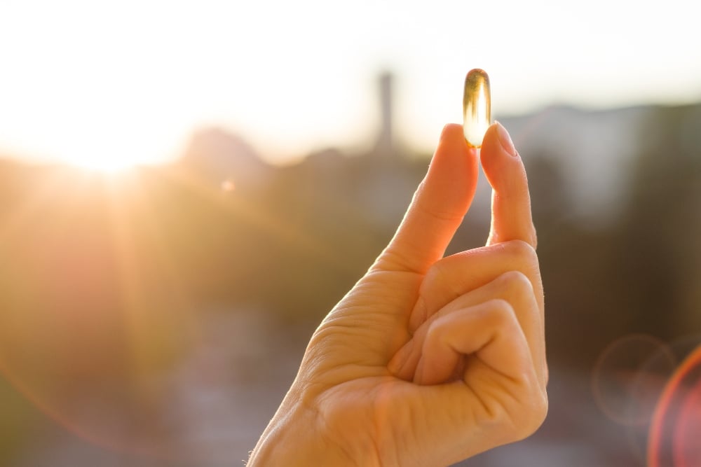 A person's hand holding a gold supplement capsule in the sunshine