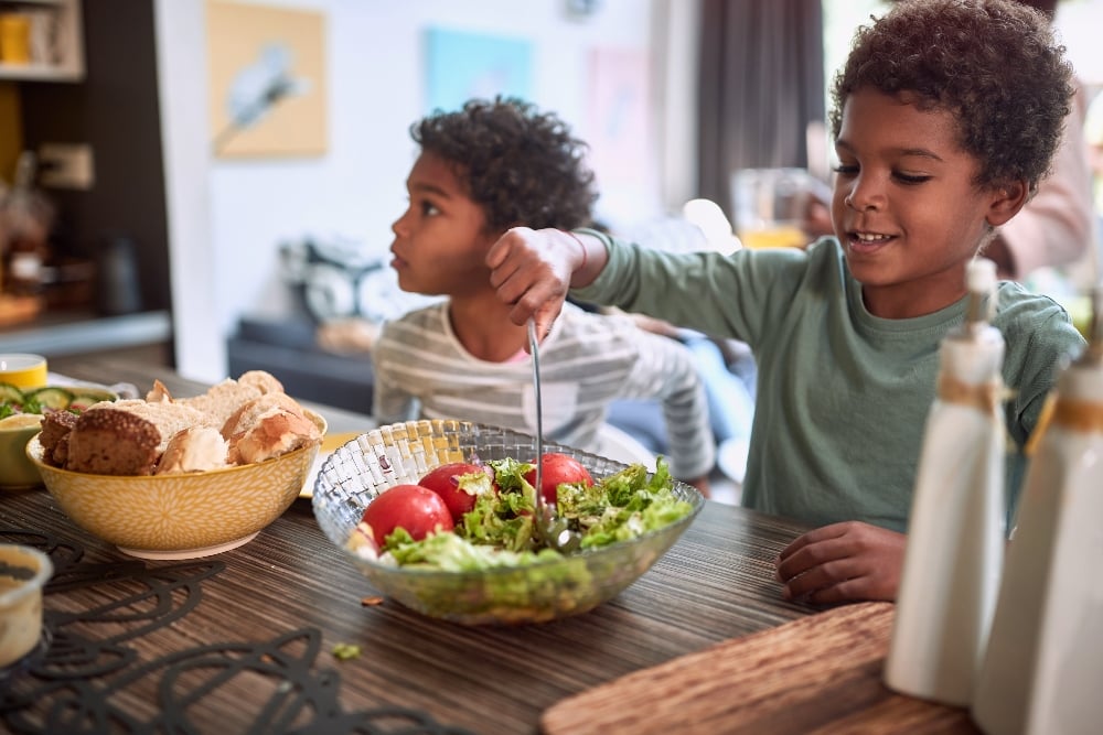 Two children sitting at a table, while one sticks a fork into some food, which can be a vegan source of vitamin D