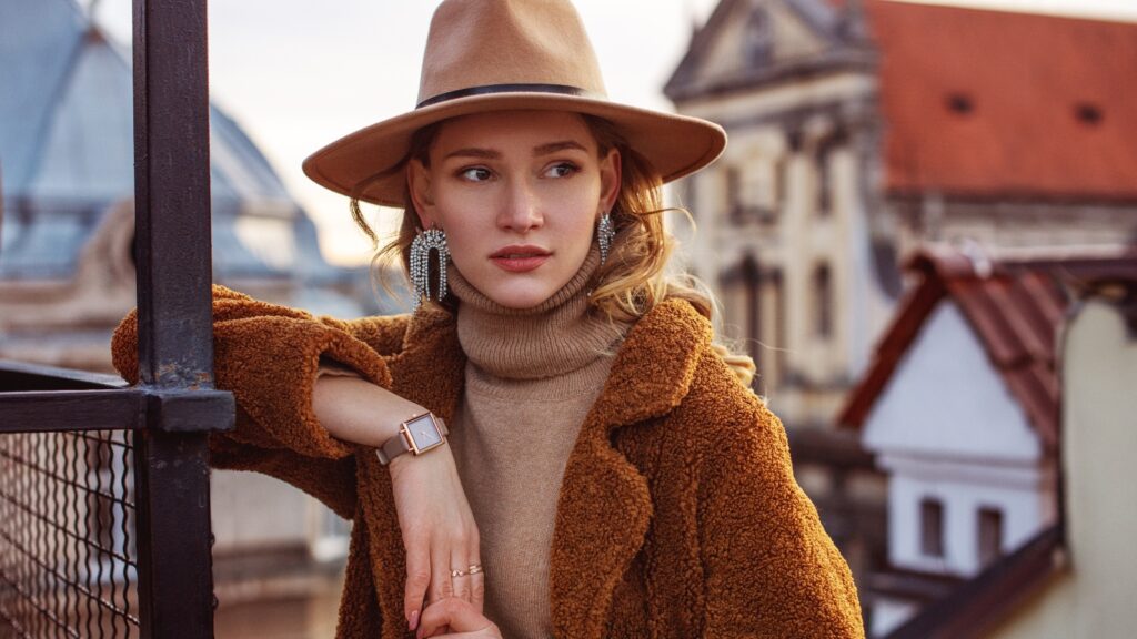 woman wearing beige hat, wrist watch, turtleneck, brown faux fur coat poses in european city