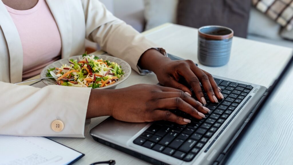 A woman eats a salad while working on her laptop