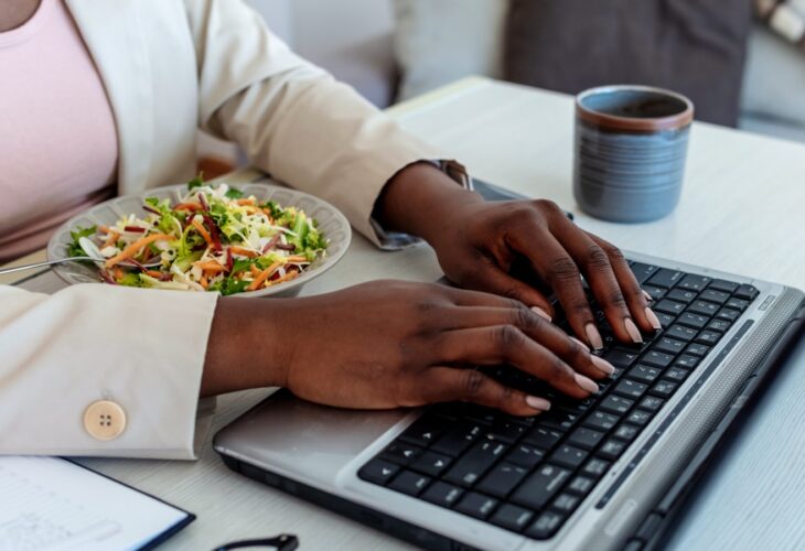 A woman eats a salad while working on her laptop