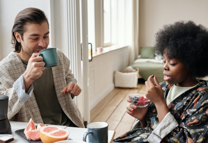 two people sit at the table drinking coffee and eating fruit