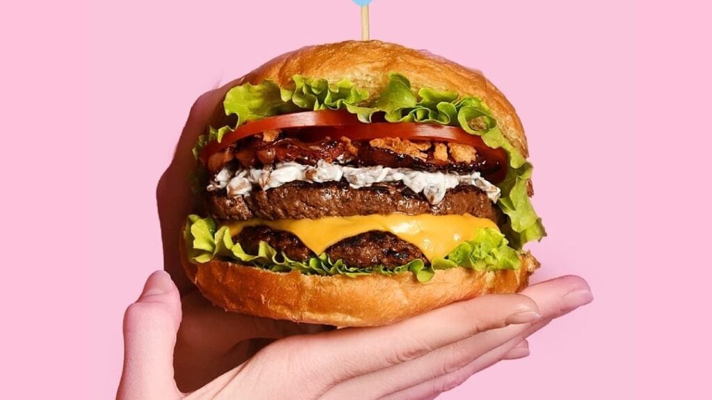 A hand holds burger in front of a pink background