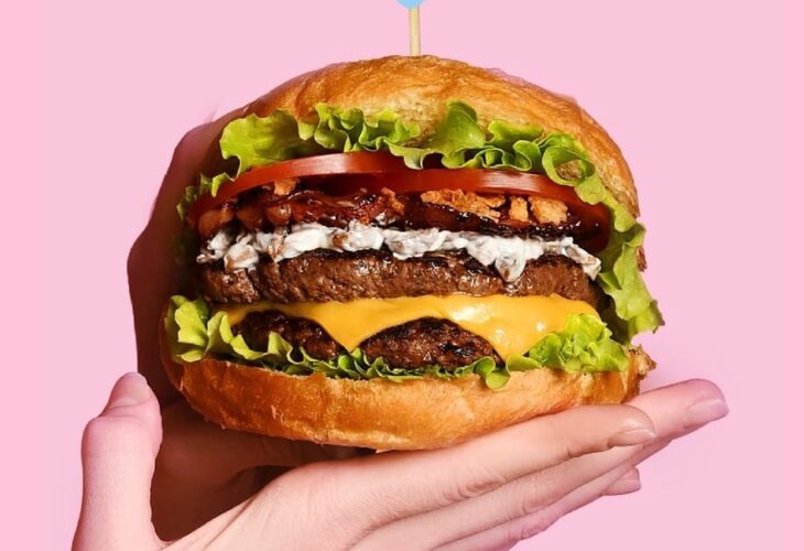 A hand holds burger in front of a pink background