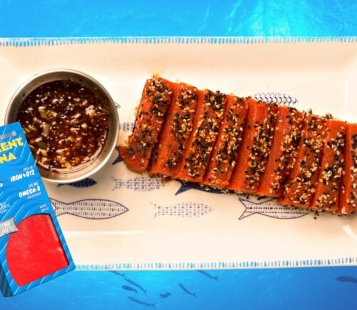Grilled vegan tuna steak on a plate beside a box of Current Foods tuna, with an ocean blue background