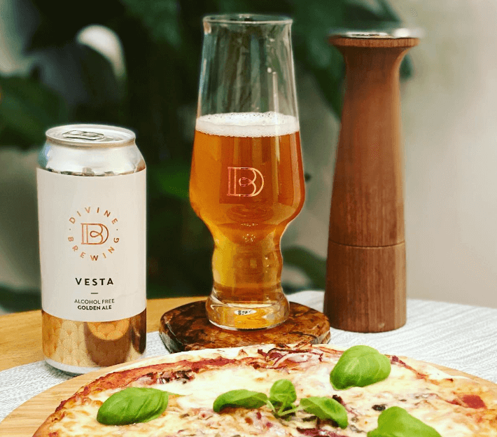 Vegan alcohol-free beer by Divine Brewing beside a cheesy pizza