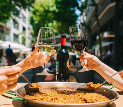 Two people clinking red and white wine glasses and eating a paella