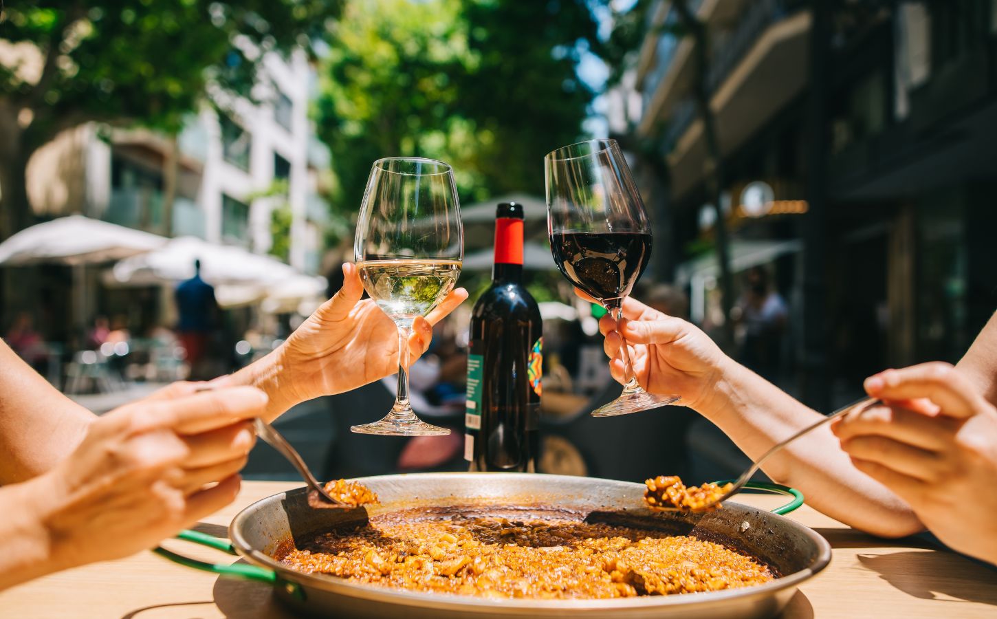 Two people clinking red and white wine glasses and eating a paella