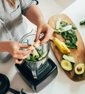 Woman preparing a healthy smoothie in a blender, with banana, green spinach, and avocado.