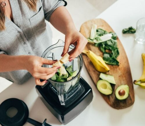 Woman preparing a healthy smoothie in a blender, with banana, green spinach, and avocado.