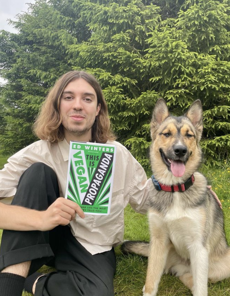 Ed Winters sits on the grass next to a dog holding his book "This Is Vegan Propaganda"
