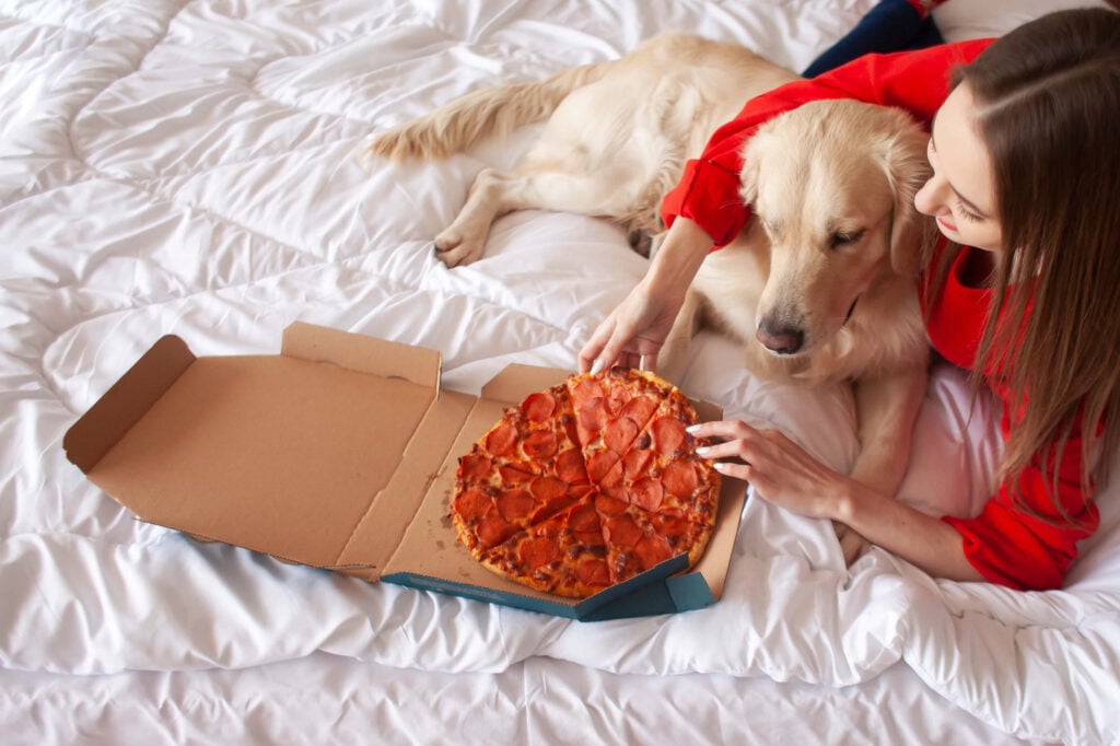 A person lying on a bed with her companion dog while eating a pepperoni pizza with animal meat