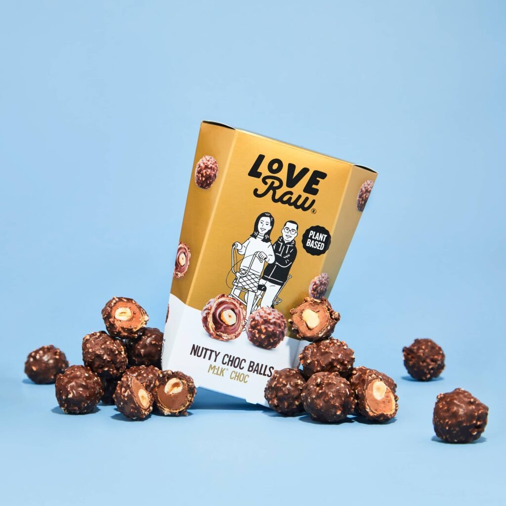 A box of vegan dairy-free Love Raw Nutty Choc Balls surrounded by unwrapped chocolate products, on a blue background