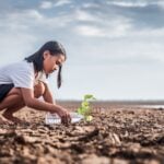 A child crouched on some barren land affected by climate change, watering a small plant with a water bottle