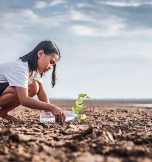 A child crouched on some barren land affected by climate change, watering a small plant with a water bottle