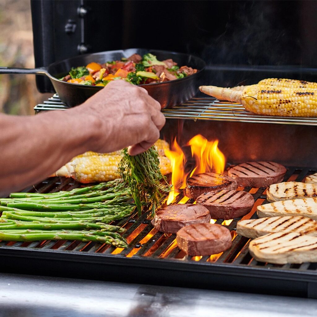 Vegan steaks on a flaming barbecue, surrounded by grilling vegetables and a hand placing more greens on the grill