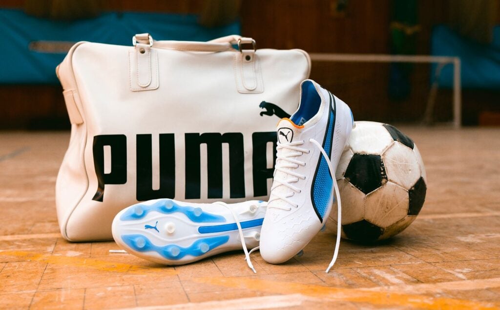 A pair of white and blue Puma vegan leather KING football boots next to a soccer ball and a Puma-branded sports bag