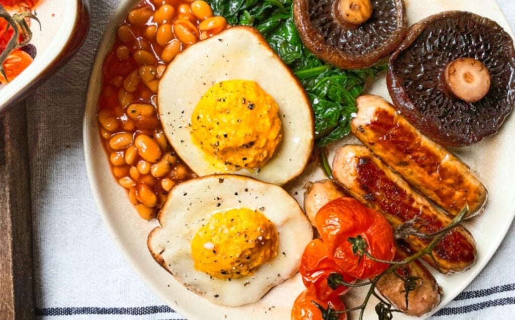 Vegan fried eggs shown as part of a plant-based fry-up on a white plate with sausages, beans and mushrooms