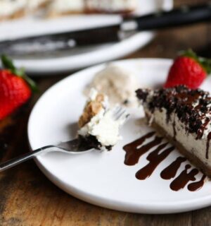 A slice of vegan Oreo vanilla cheesecake on a plate, served with strawberries, dairy-free cream and chocolate sauce