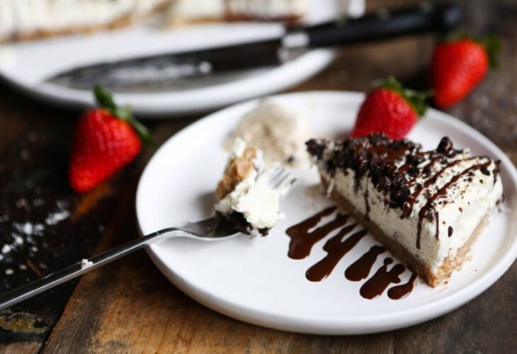 A slice of vegan Oreo vanilla cheesecake on a plate, served with strawberries, dairy-free cream and chocolate sauce