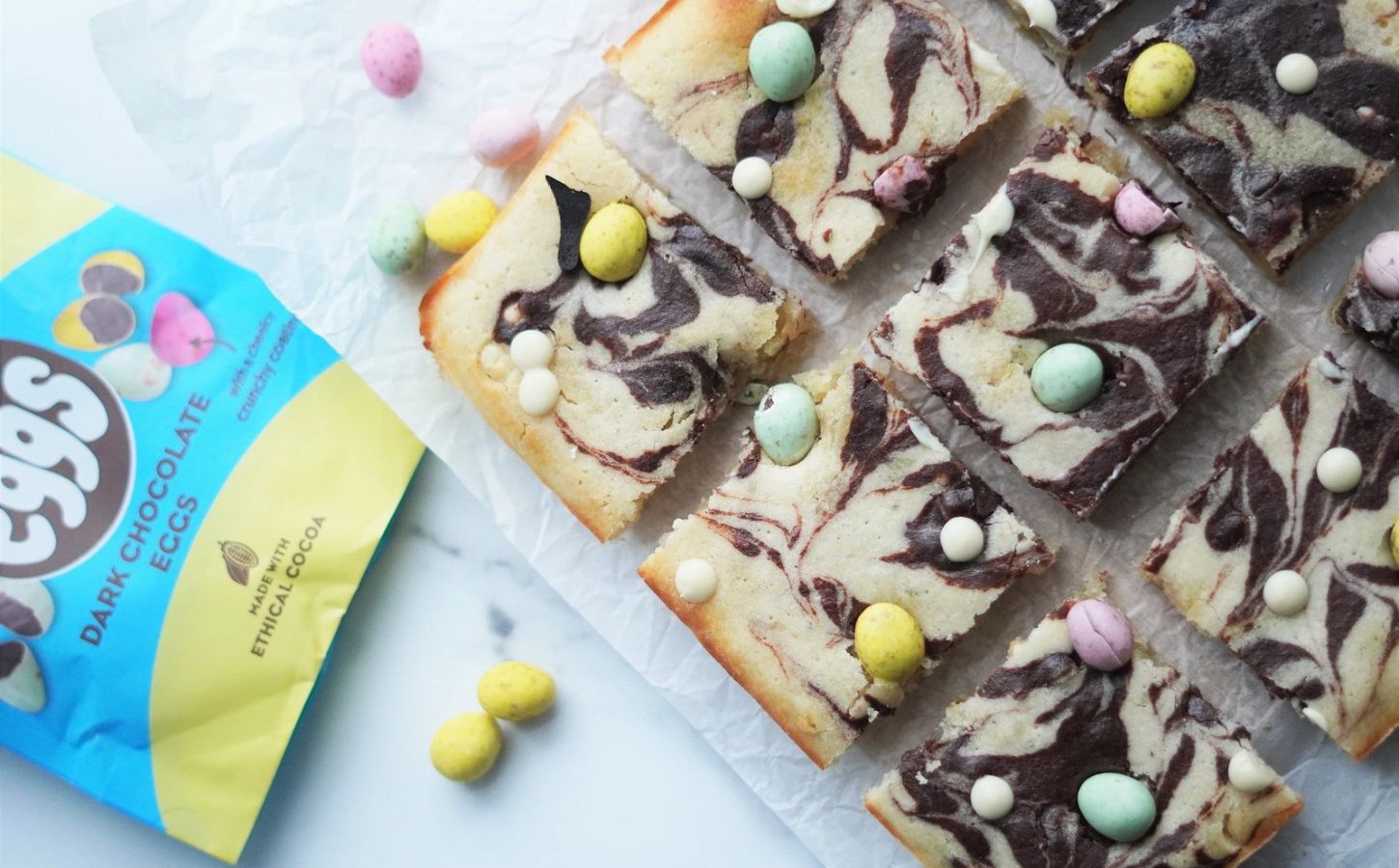 Vegan easter blondie bites laid out on a marble work surface, surrounded by vegan chocolate mini eggs