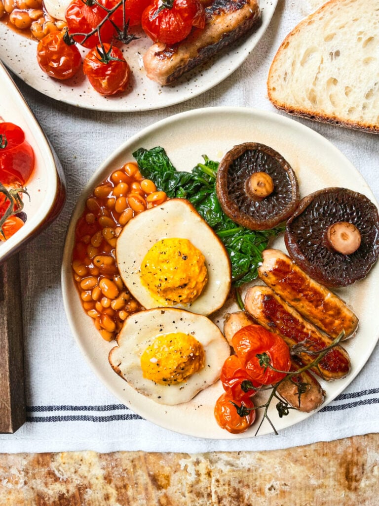 Vegan fried eggs shown as part of a plant-based fry-up on a white plate with sausages, beans and mushrooms