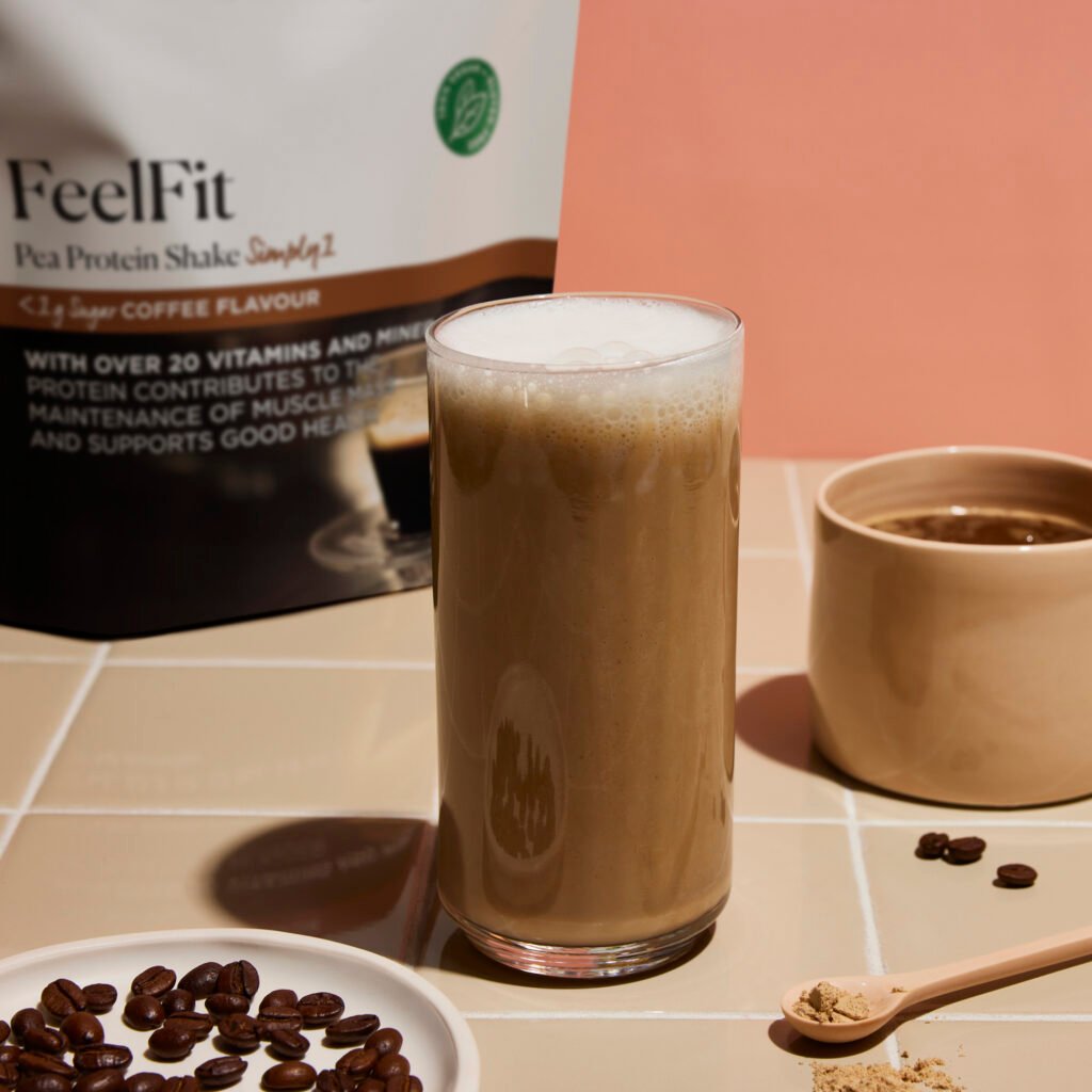 A coffee-flavoured vegan Arbonne protein shake on a tiled table surrounded by coffee beans and a protein powder packet